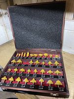 Wooden case open showing 34 yellow coated carbide tipped router bits arranged in rows with labeled slots; foam lining inside lid.