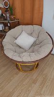 Front view of rattan papasan chair featuring large beige tufted cushion and smaller beige throw pillow.