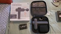 Opened box showing Sharper Image Power Percussion massager, charging cord, carrying case, and manual on a surface
