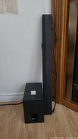 Sony black subwoofer and soundbar side by side on floor against white wall and wood cabinet.