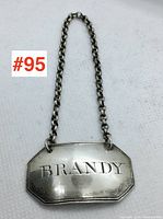 Front view of silver decanter label with 'BRANDY' engraving and attached chain laid on white fabric.