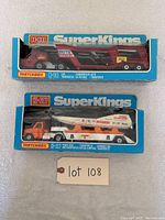 Two Matchbox SuperKings toy trucks in original boxes on white cloth surface with lot number tag.