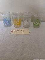 Front view of four Welch's collectible glasses with different colored fruit and character designs lined up in a row on a white surface.