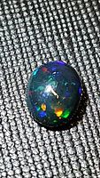 Close-up image of shiny oval cabochon black opal with vivid blue, green, and orange flashes on dark fabric background.