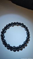 Photo showing a round stretchy bracelet made of smooth, spherical, dark metallic gray hematite beads alternating or mixed with polished black onyx beads, laid flat in a circle on a plain background with some shadowing.