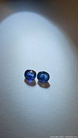 Two oval cut natural blue sapphire gemstones shown from above on white surface with light reflection, showing color and inclusions.