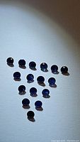 16 natural blue sapphires with diamond cut facets laid out on white background showing size and color variation.