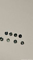 Eight small round cut sapphires arranged on a neutral background showing the teal blue to green color variations.