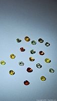 Multiple clear photos show 18 small faceted round sapphires in a range of rainbow colors arranged on a white background under bright light.