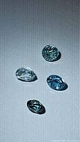 Four natural mined zircon gemstones of varying light blue to green hues on a neutral background, showing shape and facets.