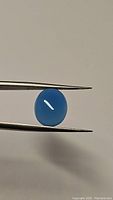 Oval lagoon blue chalcedony cabochon held by tweezers showing color and clarity against light background.