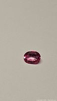 Close-up image of natural hot pink oval cut topaz gemstone against a neutral background, showing the stone's shape and color.