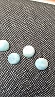 Four natural Larimar round cabochon gemstones placed on black textured fabric. Light aqua blue color with white patterning visible on three stones, one slightly paler.