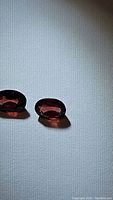 Two natural oval cut Mozambique garnet gemstones shown with visible facets, deep reddish-purple color, and reflection indicating clarity.