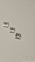 Three square cut natural rose quartz gemstones placed on a plain surface, showing size and clarity.