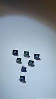 Seven princess cut blue teal sapphires arranged in a pattern, showing color and cut clearly on a white background.