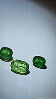 Three natural tsavorite garnet gemstones displayed on white background under light showing rich green color and some inclusions.
