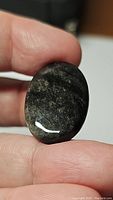 Close-up view of the polished oval cabochon of silver sheen obsidian held between fingers showing reflective silver sheen and dark base.