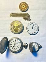 Antique silver pocket watches, pocket watch chain, and miscellaneous watch parts laid on white background.