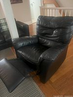 Black leather recliner chair extended with leg rest visible, placed on area rug over hardwood floor, next to a black sofa.