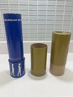 Blue kaleidoscope tube standing with two cylindrical cardboard tubes behind, all on a white surface with tiled grey background.