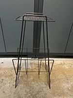 Black metal mid-century modern magazine rack with table top, standing on a concrete floor, photographed from the front against a blue background.