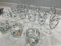 Top angle showing full set of 20 glasses arranged on a surface, mixing stemmed and tumbler types with atomic amoeba and boomerang designs.
