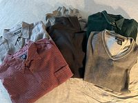 Photo showing red checkered shirt, olive green ribbed polo shirt, light brown V-neck sweater, and other folded tops.