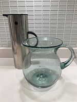 Side view showing the stainless steel Stelton coffee jug with black handle and clear hand blown glass sangria jug with bulbous body and single handle.
