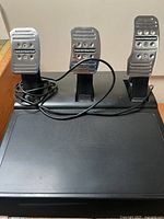 Top view of three metallic racing pedals mounted on a black base with visible Thrustmaster branding and attached cables.