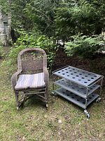 Photo of rustic wicker rocking chair and metal multi-tier side table on grass outdoors, showing full view of both items.