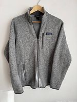 Full front view of the men's grey Patagonia fleece jacket hung on wooden hanger against white background. Shows full jacket and pocket zipper details.