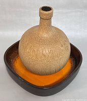 Front angle showing the round textured beige stoneware vase placed inside the dark brown and orange square ceramic bowl.