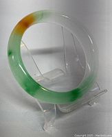 Natural jade bangle displayed on clear stand showing its D-shaped cross-section and jade's green, white, and brown coloring.