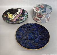 Three items shown: cloisonne plate with dark blue and gold floral design, and two-piece stackable porcelain box with colorful iris floral motifs and geometric patterns.