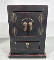 Front view of vintage Asian-style black wooden jewelry cabinet with double doors and single bottom drawer featuring bronze-tone hardware and character motif pull rings.