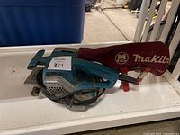 Blue and black Makita belt sander with power cord and red dust collection bag on a white table.