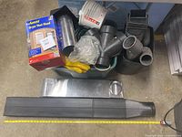 Top view of assorted metal and plastic dryer vent duct pieces, fittings, gloves in green plastic tub, and a boxed dryer vent hood.