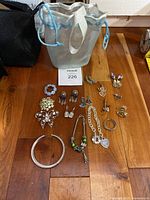 Photo shows multiple pieces of assorted rhinestone and metal jewelry including brooches, earrings, charm bracelet, and necklaces arranged on a wooden surface next to a silver cloth bag