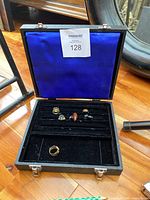 Open view of black jewellery box showing blue interior lining and six assorted rings inside on black velvet slots. One gold-tone ring outside the slot area.