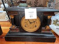 Front view of the black wooden mantel clock with ornate gold-tone dial and two columns on each side.