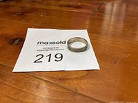 Silver band ring lying on a wooden surface next to a numbered paper with the lot number 219.