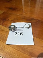 Two rings on paper showing lot number; one large with clear rectangular gem and ornate silver band, second smaller with green and clear gems on a silver band.