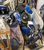 Two push golf carts with wheels, golf clubs in bag, blue golf umbrella visible among items, hockey bags and other equipment visible in background.
