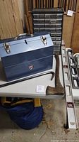 Blue metal toolbox, small drawer organizer, axe, crowbar, and levels on a table in a garage.