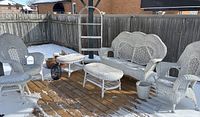Wide shot of white wicker patio set on wooden deck with some snow, showing loveseat, two chairs, two tables, waste basket, plant pots, and black lantern.