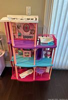 Full front view of the Barbie Dream House with three levels showing colored floors, furniture items, and printed backdrops.