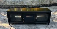 Front view of black wooden credenza with three compartments and glass panel doors on a concrete surface outdoors.