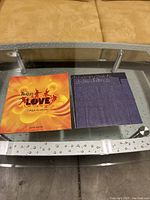 Top view of two booklets side by side on glass coffee table; one is Beatles Love bright orange cover booklet, the other purple textured booklet.