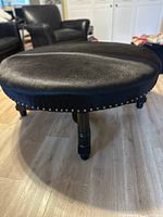 Front-side view of round black cowhide ottoman showing top upholstery and nailhead trim